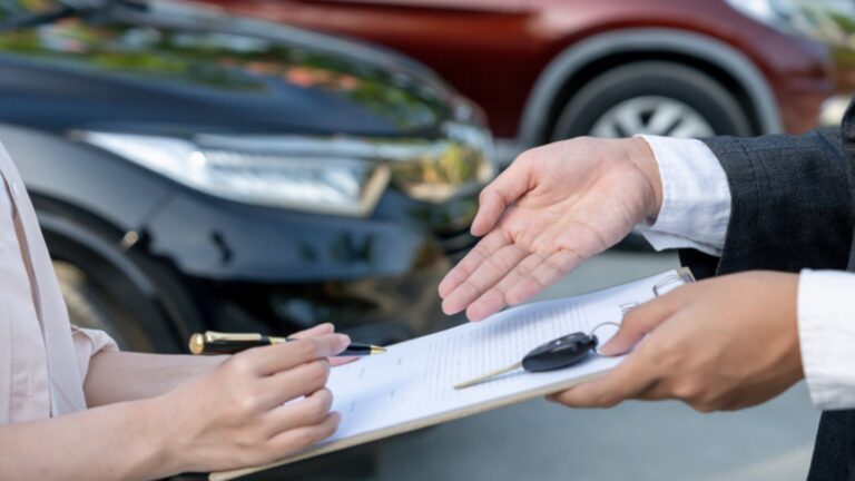 A buyer holds a pen over a sales contract and car key near parked cars