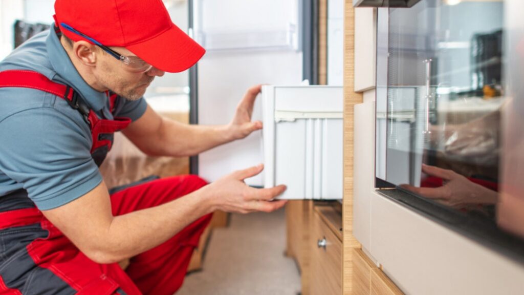 A technician in a red cap removes a refrigerator drawer inside a motorhome kitchen