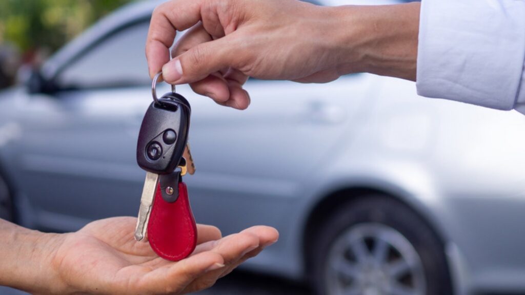 Car key with a red leather tag passed into an open hand beside a silver car